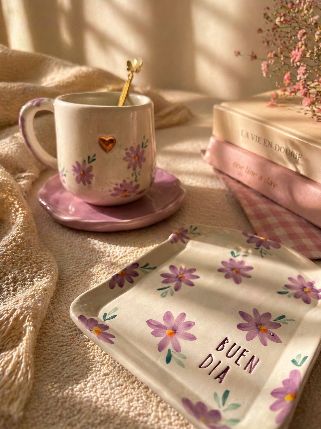 Taza floral y bandeja a juego sobre un mantel rosa bajo la cálida luz del sol, con una cuchara dorada y una caja de regalo cerca.