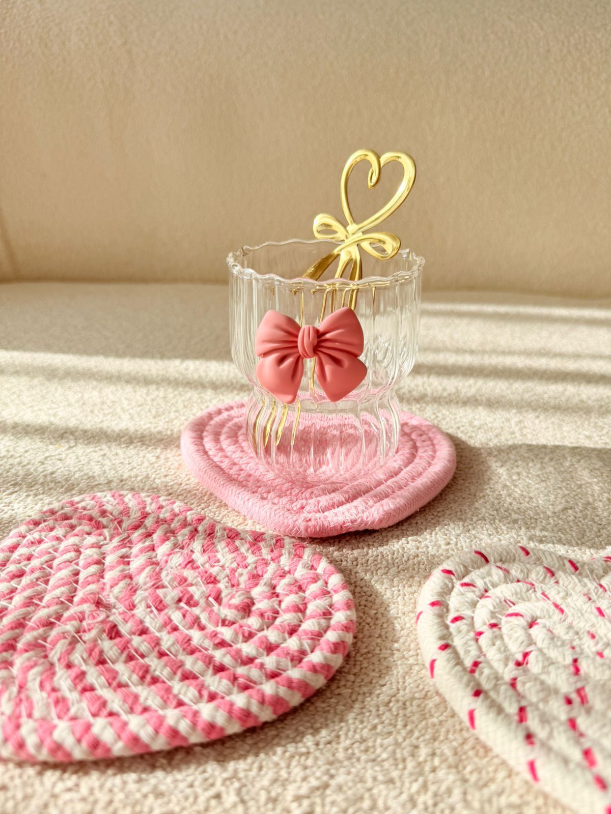 Manteles individuales rosas tejidos y un vaso transparente con un lazo color coral, dispuestos sobre un sofá beige a la luz del sol.