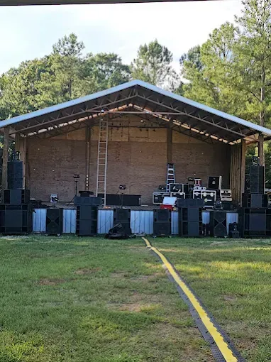 Outdoor stage setup with sound equipment. Green grass and trees in the background. A yellow cable lies in the foreground.