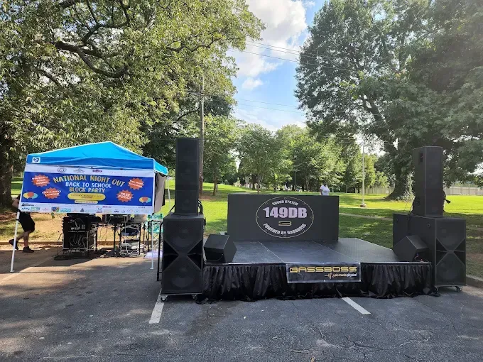 Outdoor stage setup with sound equipment, tent, and backdrop under a blue sky, in a park.