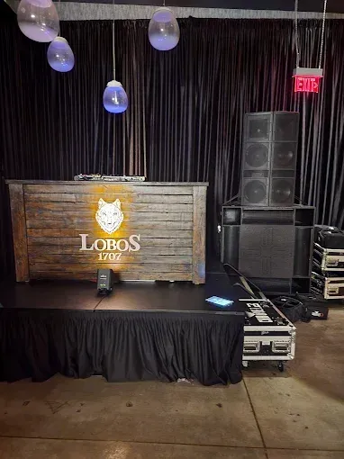 DJ stage with sound equipment. Wooden bar with a wolf logo, black stage skirt, dark background, and hanging lights.