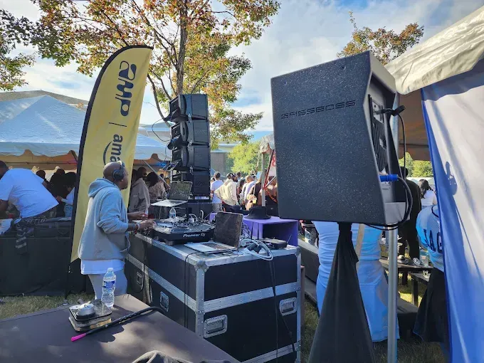 DJ equipment set up outdoors at an event, including speakers, turntables, and a sound system. People in the background.