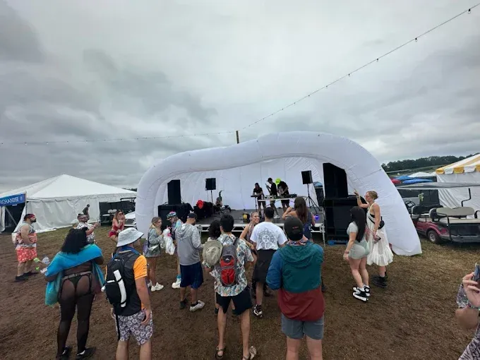People dance in front of a white stage at an outdoor music festival on a cloudy day.