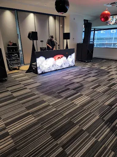 DJ at a party, behind a table with LED screen, surrounded by speakers. Room with patterned carpet.