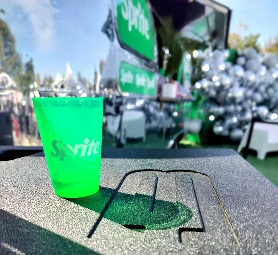 Green Sprite cup on a textured surface, with a food stall in the background.