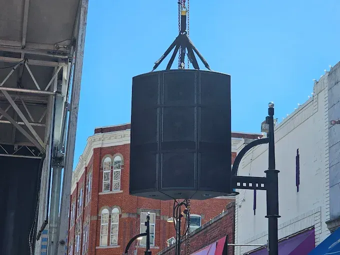 Large black speaker system suspended above a street, set against a blue sky and brick buildings.