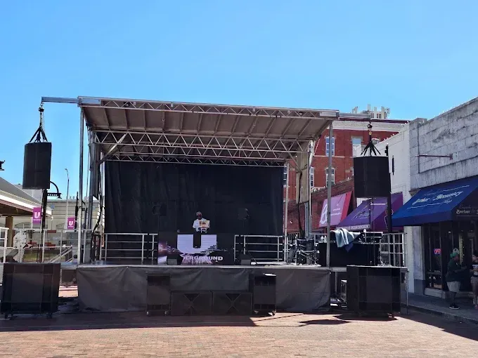 A DJ performs on an outdoor stage, set up in a town square. Speakers and equipment are arranged, blue sky above.
