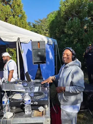 DJ at outdoor event, wearing headphones. DJ equipment and a speaker visible. Blue tent and trees in background.
