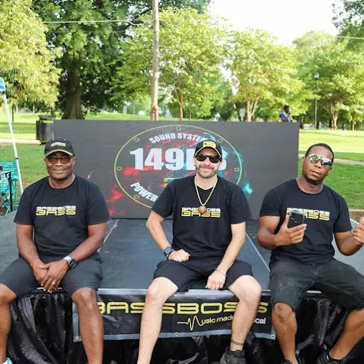 Three men in black shirts sit in front of a stage with a sound system backdrop in a park.