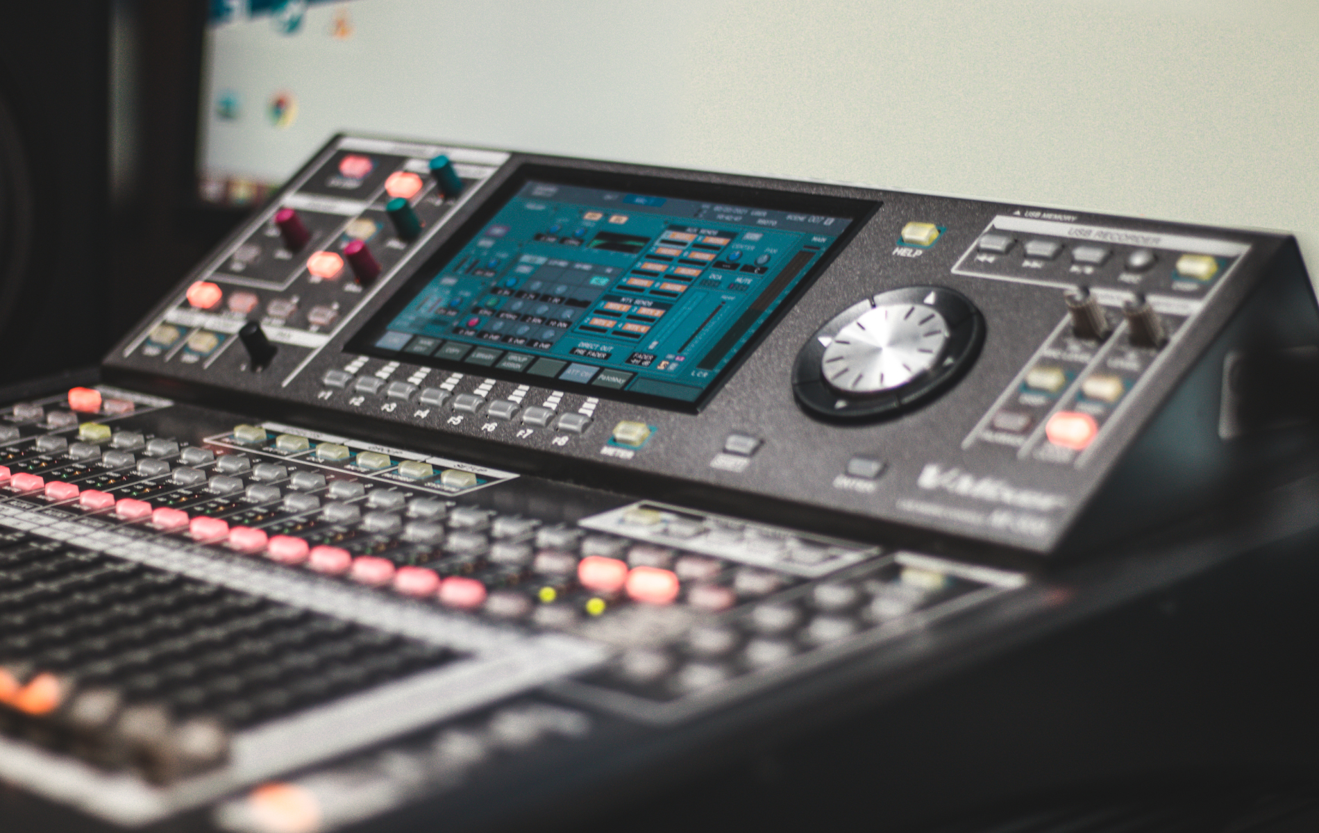 Audio mixing console with lit buttons, display screen showing audio levels, and various knobs and faders.