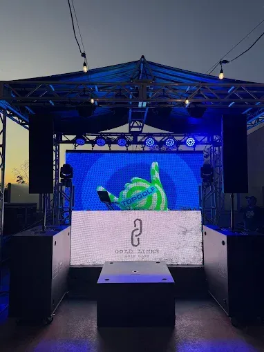 DJ stage setup, outdoor. LED screens with blue and green graphics, truss lighting, speakers, and a DJ booth.