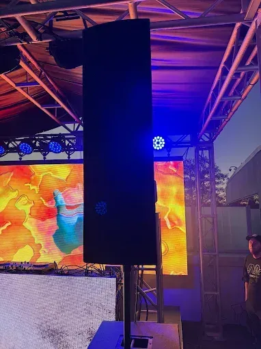 Tall black speaker on a stand in front of a colorful stage screen, outdoor setting.