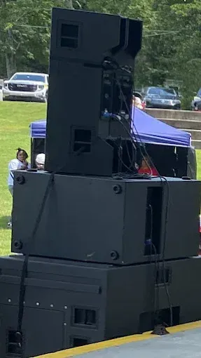 Black speaker system stacked outdoors, with a person and tent in background.