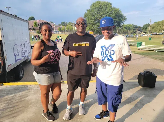 Three people standing outside. Woman in shorts, man in black shirt, man in blue pants. Outdoors, sunny.