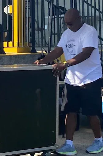 Man in white shirt and shorts lifting a dark box outdoors near a yellow structure.