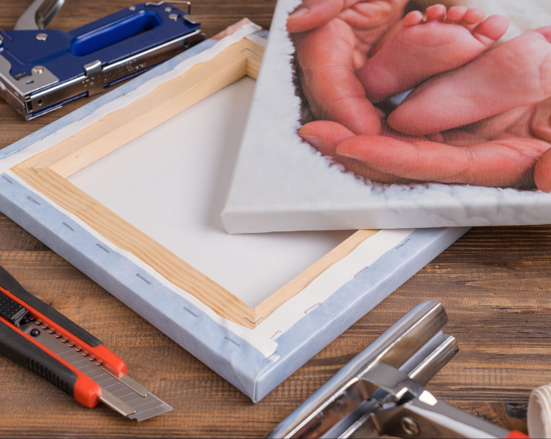 Canvas on a wooden table with art supplies: stapler, utility knife, and a hand-painted canvas in progress.