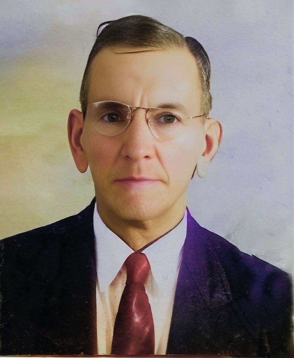 Studio portrait of a man in a dark suit, white shirt, red tie, and glasses against a light background