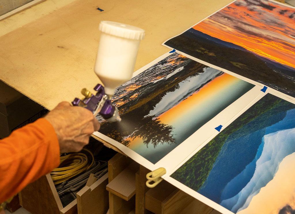 Person using a spray gun to coat a landscape photo print on a worktable