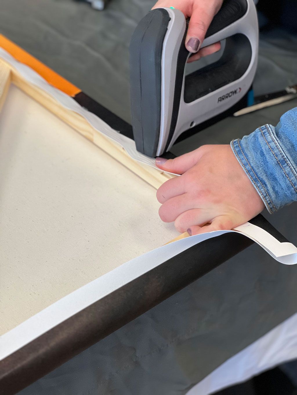 Hands using a staple gun to attach white fabric to a wooden frame on a gray surface