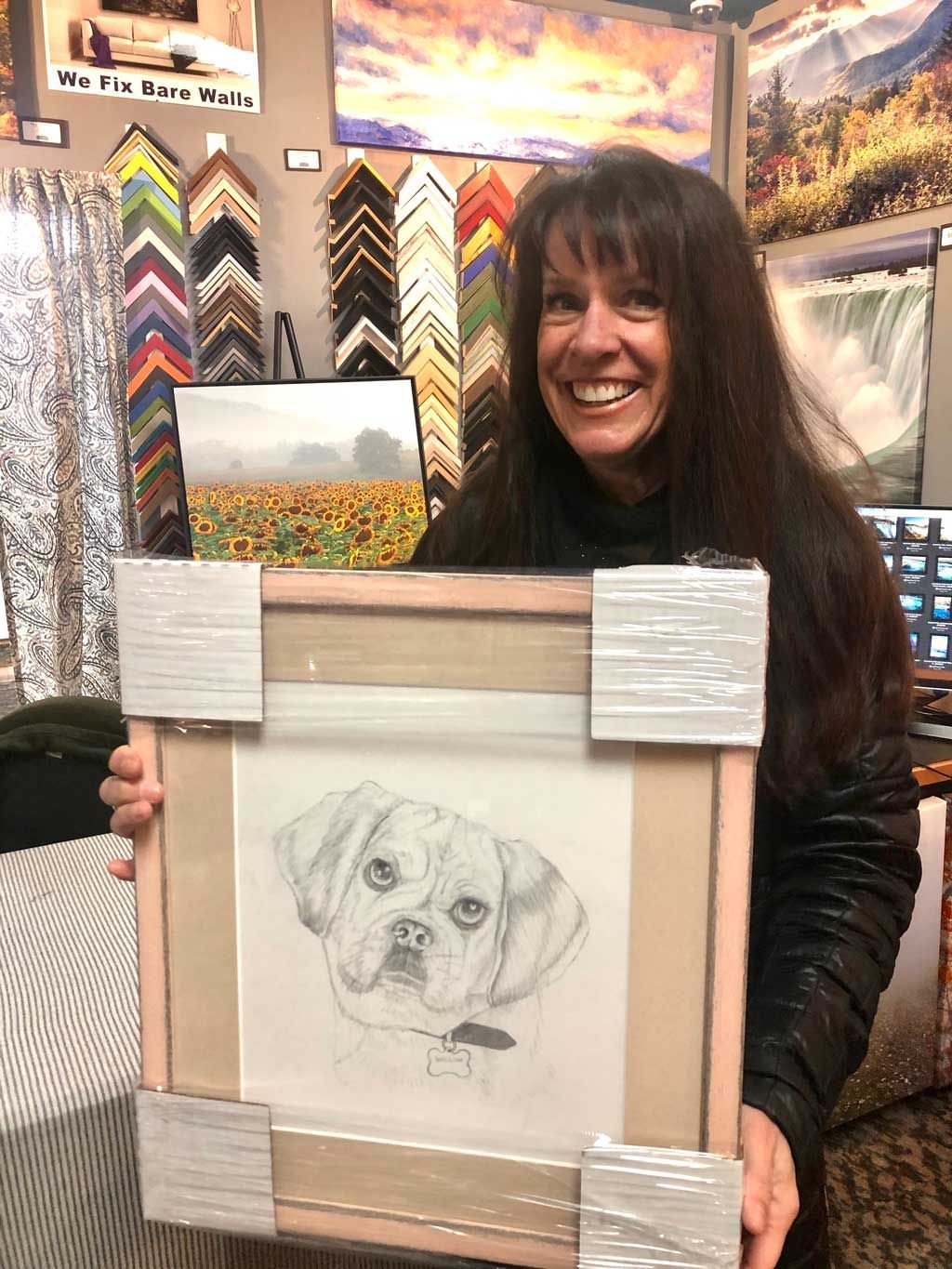 Woman smiling and holding a framed pencil sketch of a dog in an art display booth