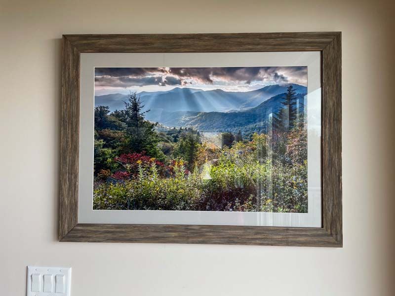 Framed landscape photograph of mountains, clouds, and wildflowers on a wall