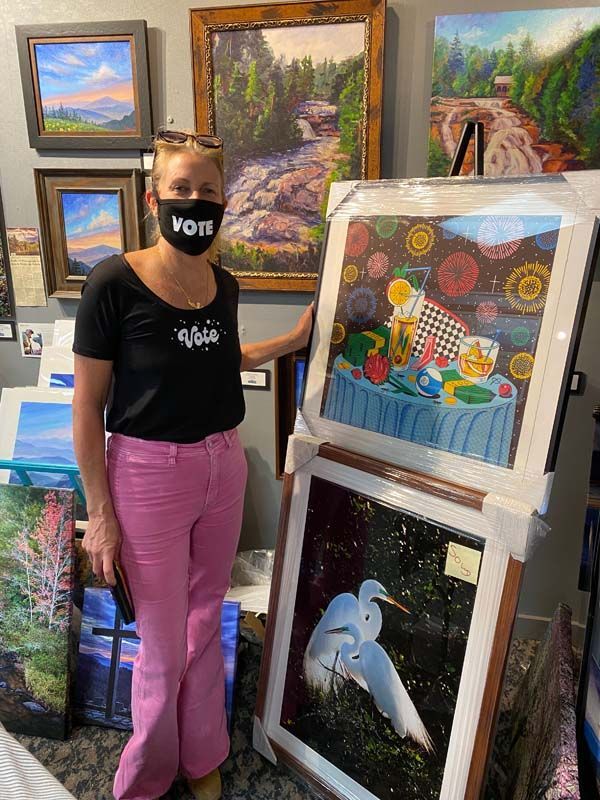 Artist stands beside colorful framed paintings in a gallery, wearing a “VOTE” mask and pink pants.
