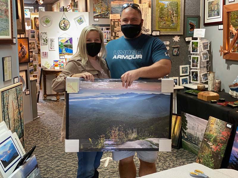 Two masked people in an art gallery holding a landscape print, surrounded by framed paintings.