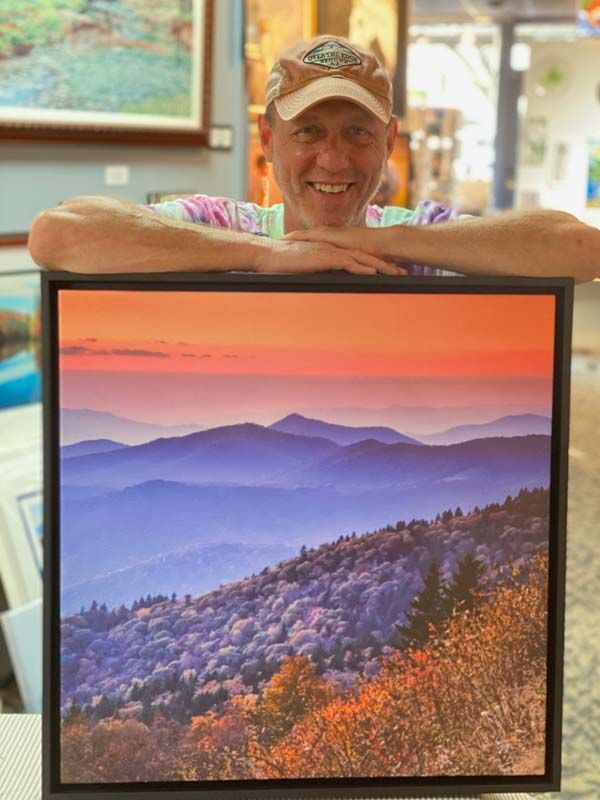 Smiling man leaning on a framed sunset mountain landscape print in a bright room