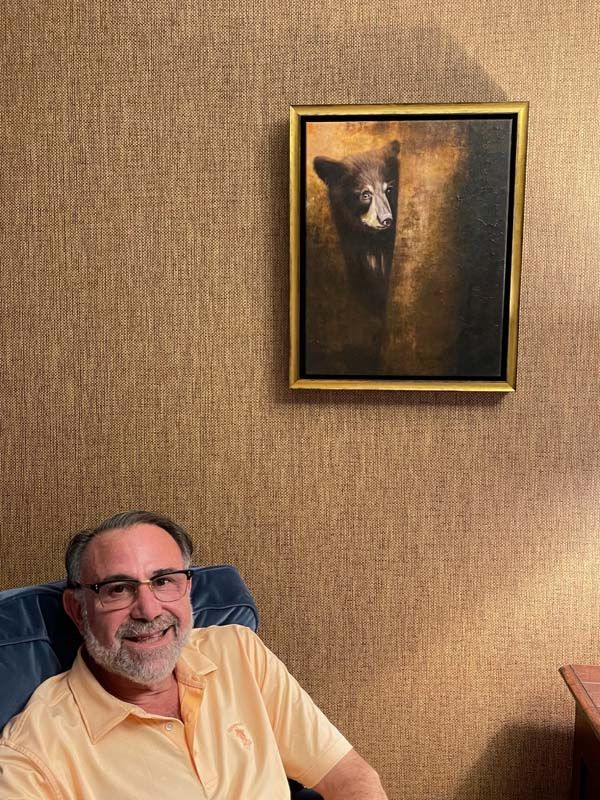 Smiling man seated beside a framed painting on a textured tan wall
