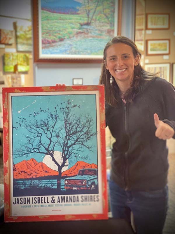 Woman holding framed poster with a tree silhouette and red border, indoors, smiling at the camera