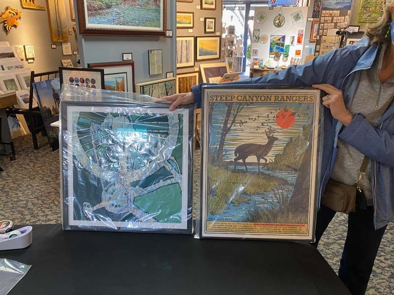 Person holding two framed art prints in a gallery, one green abstract and one with a bird design.