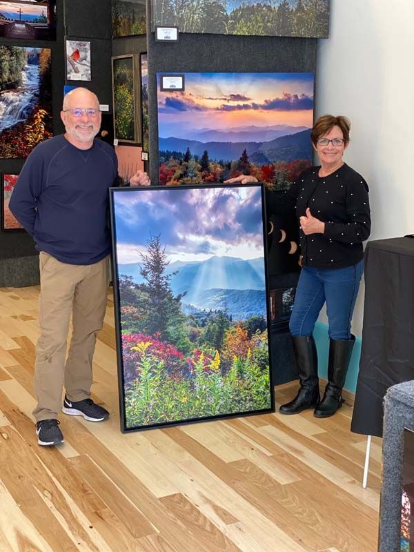 Two people pose indoors beside framed landscape paintings on display.