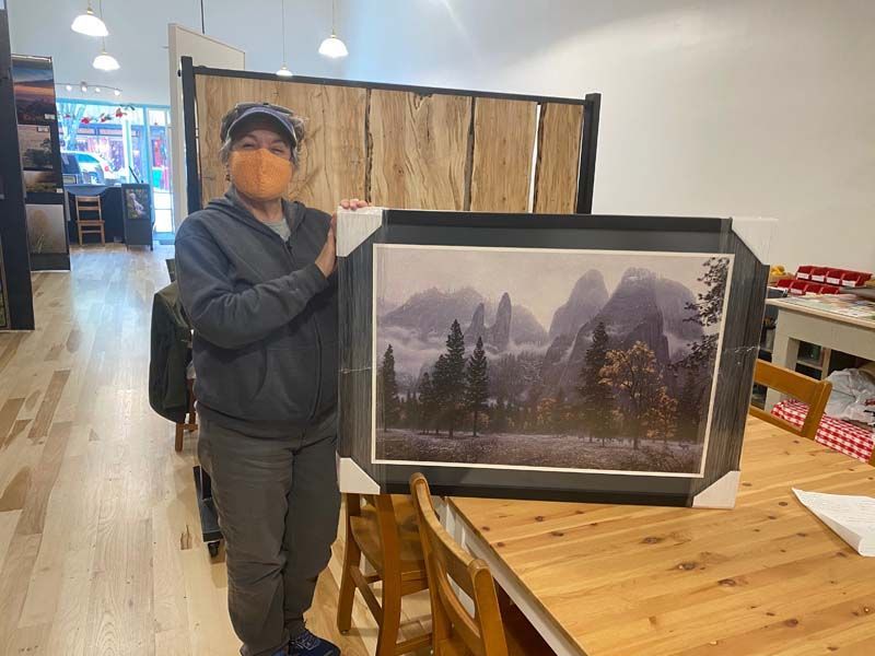 Person holding framed landscape print beside a wooden table in a store or gallery.
