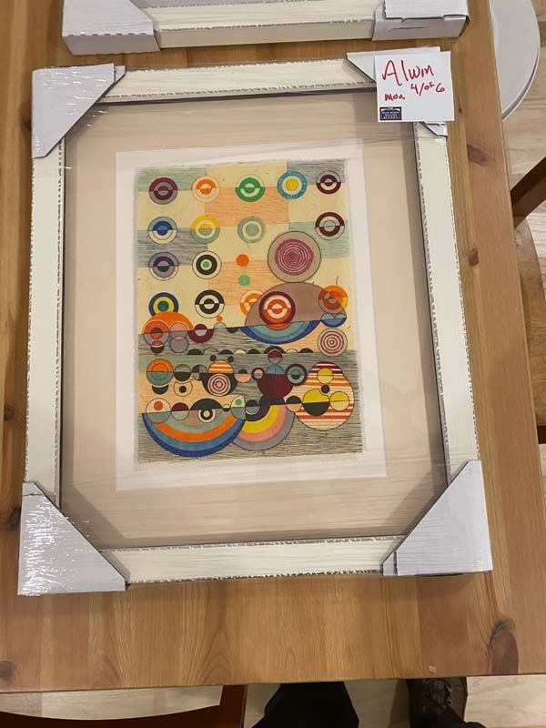 Framed abstract artwork with colorful circular patterns, wrapped in cardboard corners on a wooden table.