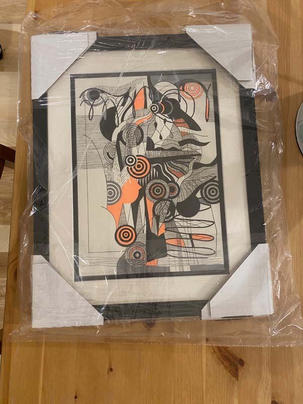 Abstract black-and-orange framed artwork wrapped in plastic on a wooden floor