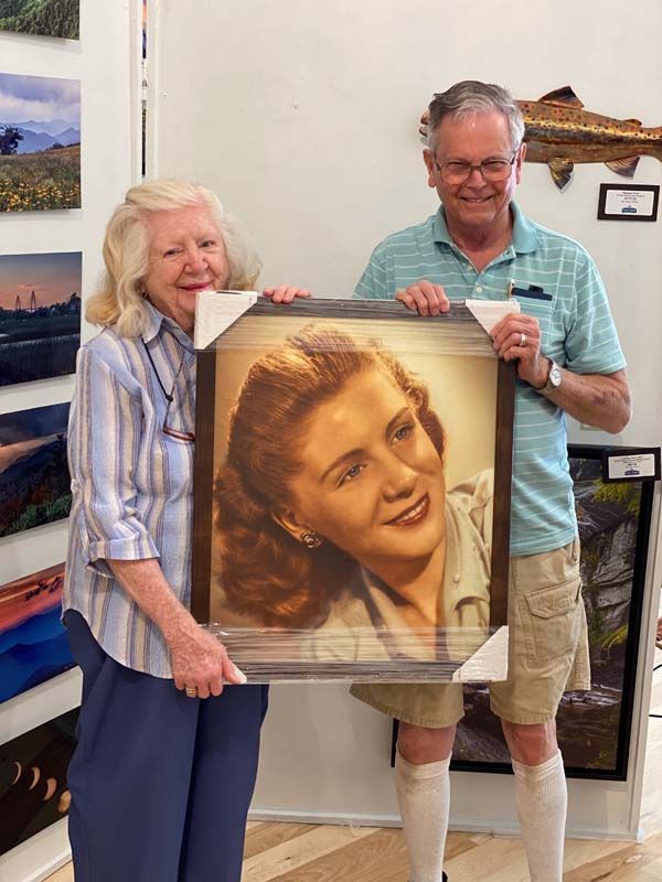 Two people holding a framed portrait in a gallery, smiling beside wall-mounted landscape photos.