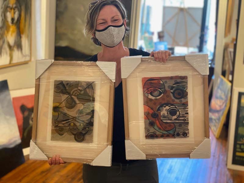 Masked person holding two framed abstract artworks indoors