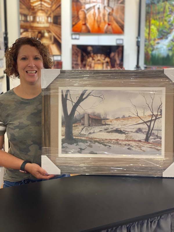 Person holding a framed winter landscape print at a display table in an art gallery