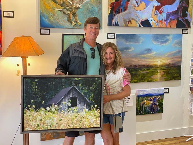 Two people holding a framed painting in a gallery with colorful landscape artwork on the walls