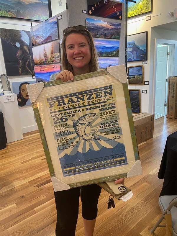 Woman holding a large vintage-style poster in a bright gallery with framed landscape artwork on the walls