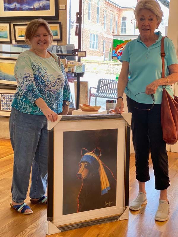Two women standing beside a framed painting in an art gallery, with windows and artwork in the background.