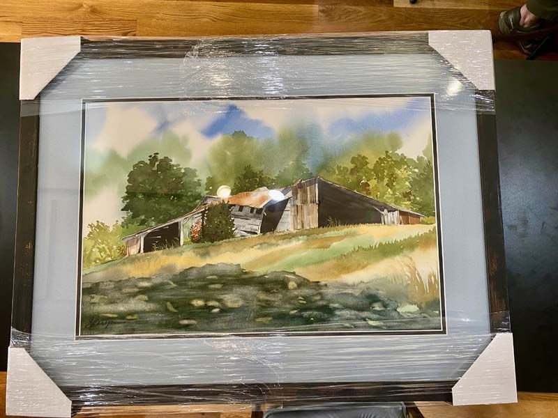 Framed watercolor of a house and trees beside water, wrapped in plastic on a wooden table.