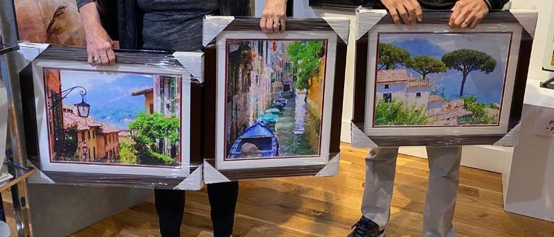 Three people hold framed paintings of a colorful street, garden gate, and lakeside landscape.