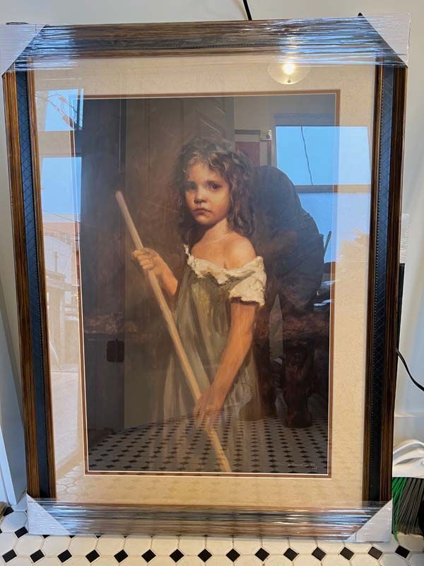 Framed portrait of a child in a white dress holding a broom, behind glass with a reflective glare