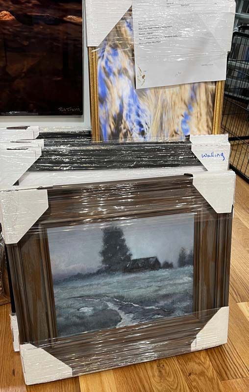 Stacked framed landscape paintings in a store display, with brown and blue-toned artwork in plastic wrap.