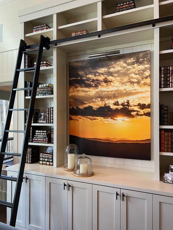 Built-in bookshelf with ladder and framed sunset landscape above cabinets