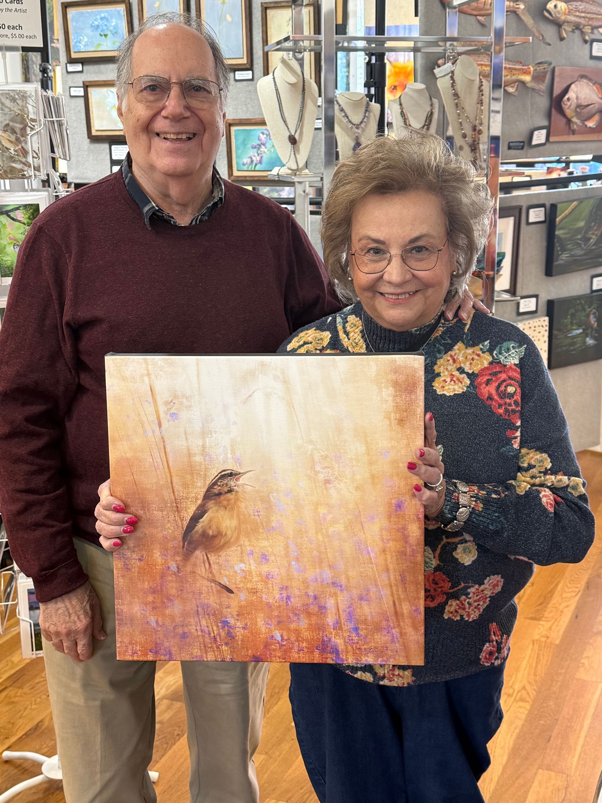 Two people smiling indoors, holding a warm-toned painting with a bird on it.
