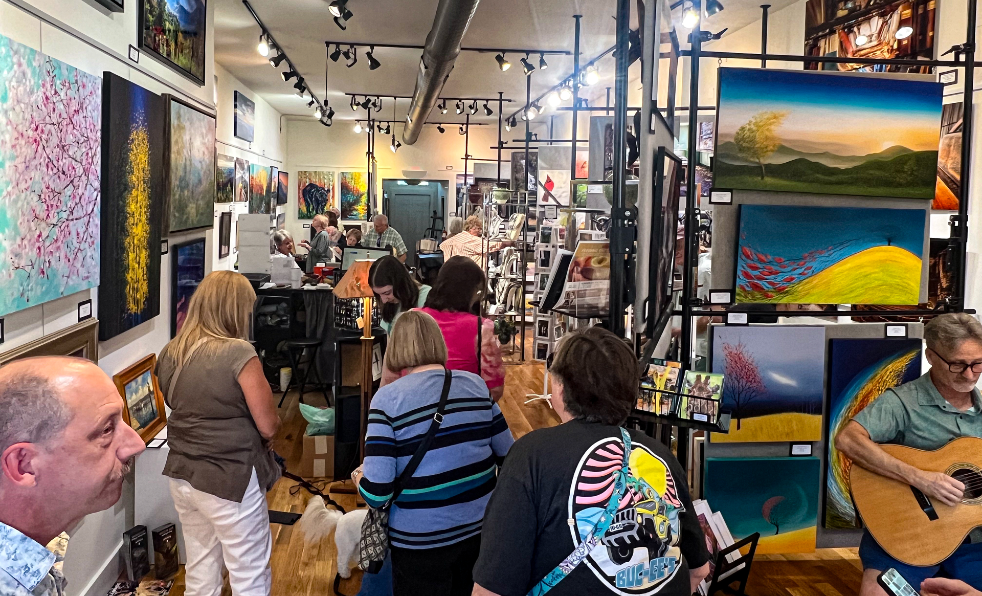 Art gallery with people browsing paintings; a musician plays guitar on the right.