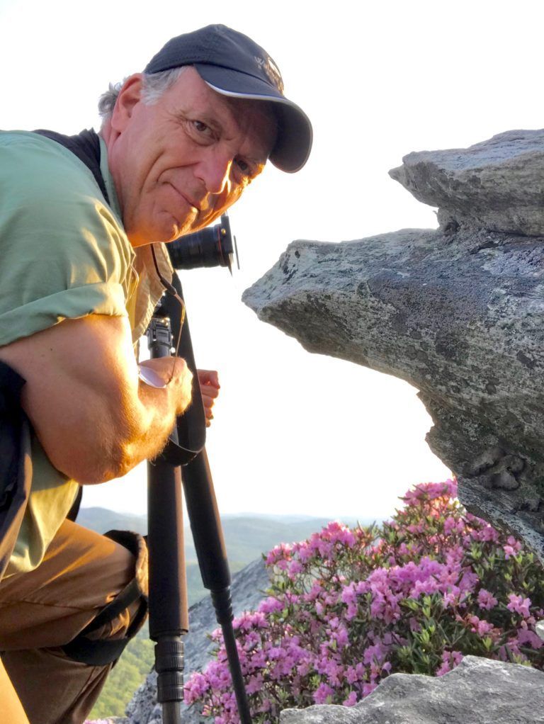 Man in a cap crouches beside a tripod on a rocky cliff with pink flowers and a hazy mountain view