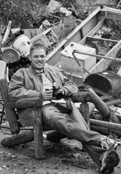 Man sitting in a chair holding a camera, with scattered tools and equipment behind him.